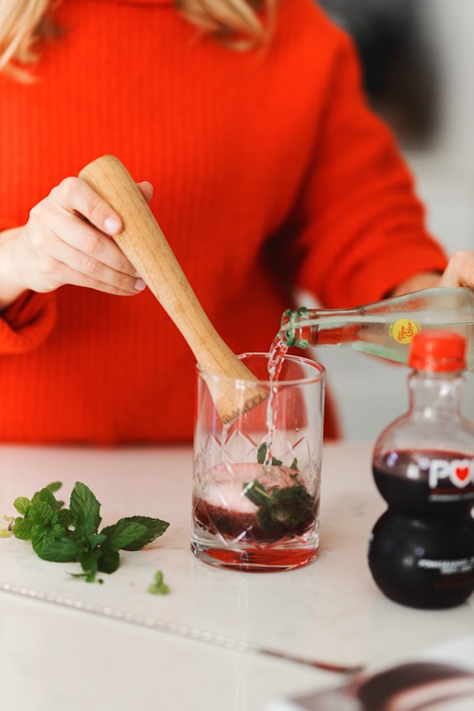 A girl in a red sweater pouring sparkling water and cranberry juice into a cocktail glass