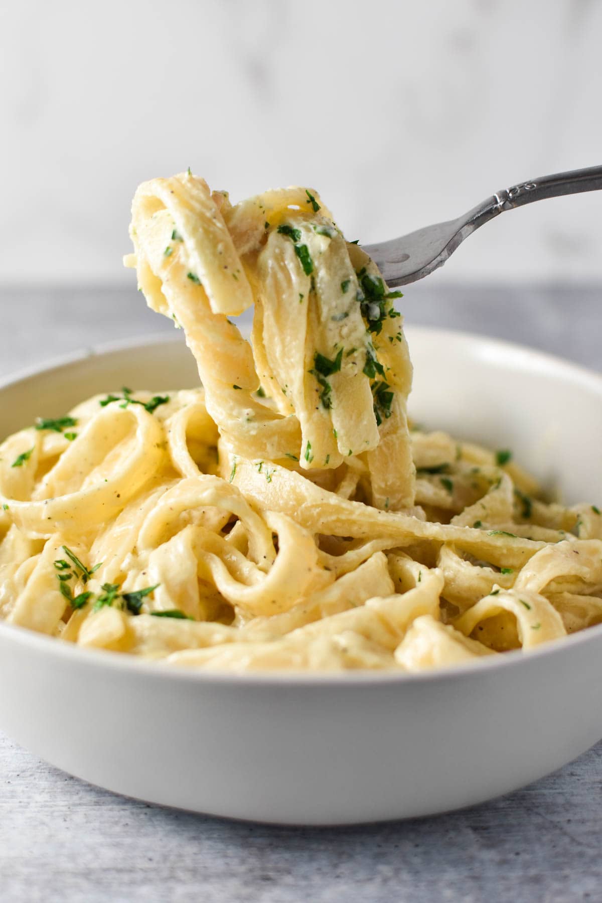 A fork holding creamy pasta thats dangling over a white bowl.
