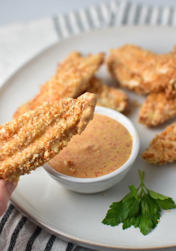 A chicken tender dipped in honey mustard sauce with a bowl of sauce in the background next to chicken tenders