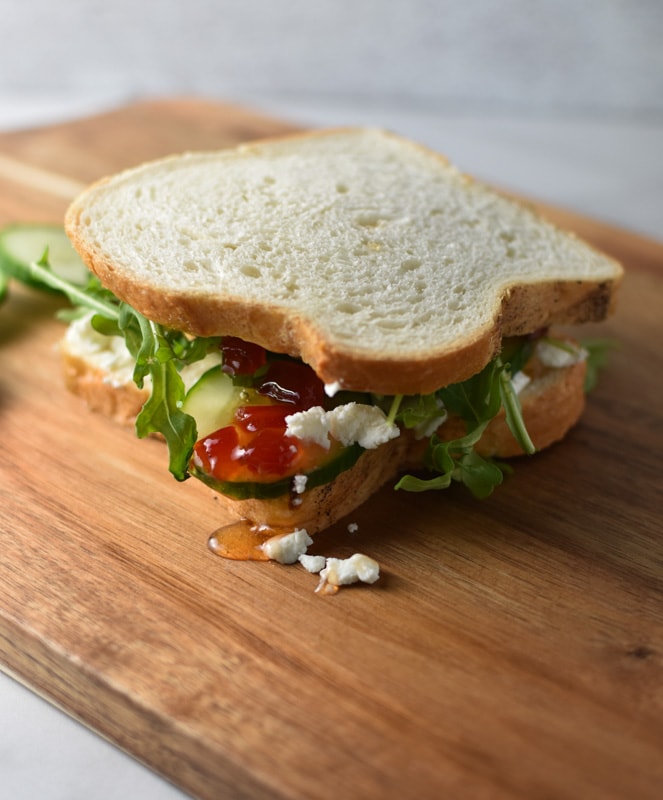 A goat cheese, chili jam, arugula, and cumber sandwich on a wooden cutting board