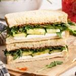 Two goat cheese sandwiches with cucumber and arugula stacked on top of each other on a cutting board.