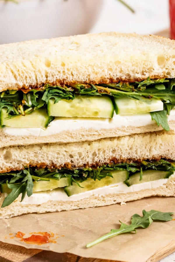Two goat cheese sandwiches with cucumber and arugula stacked on top of each other on a cutting board.