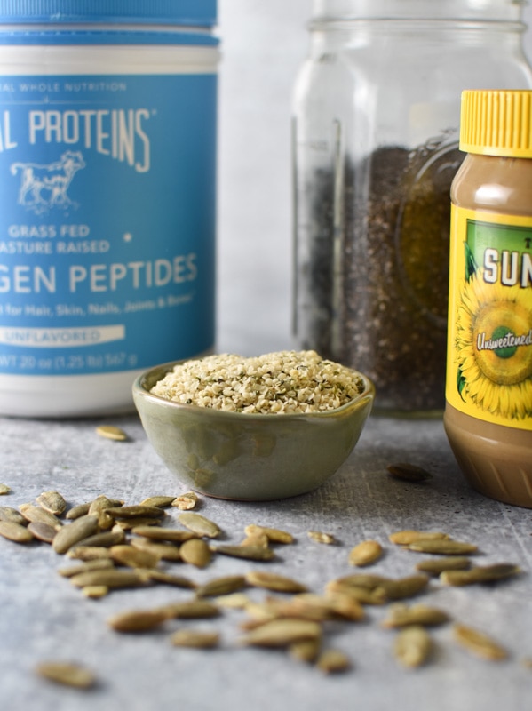 A container of collagen peptides next to chia seeds, sunbutter, pumpkin seeds, and hemp seeds on a gray surface