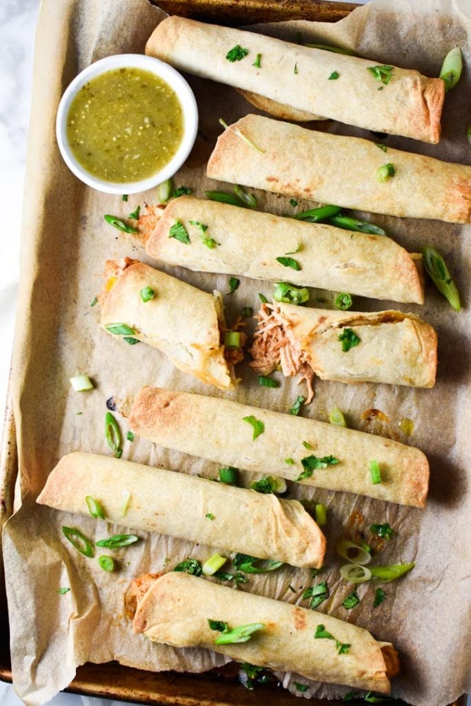 A pan of baked creamy chicken taquitos, one broken to show the filling, topped with cilantro with salsa verde on the side