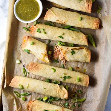 A sheet pan of baked creamy chicken taquitos, one broken to show the filling, topped with cilantro with salsa verde