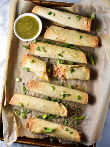 A sheet pan of baked creamy chicken taquitos, one broken to show the filling, topped with cilantro with salsa verde