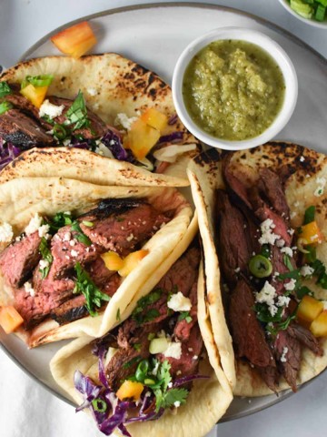 Grilled flank steak tacos lined up on a grey plate topped with cilantro and salsa verde