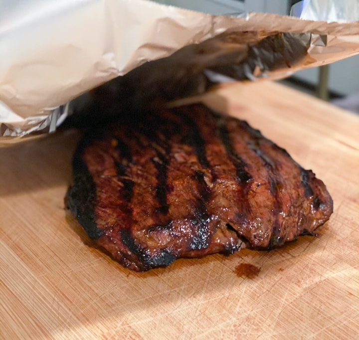 A steak that's been grilled on a wooden cutting board with a tin foil cover