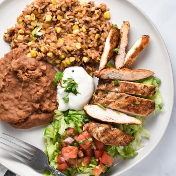 Mexican grilled chicken on a plate with beans, rice, lettuce, tomatoes, and cilantro