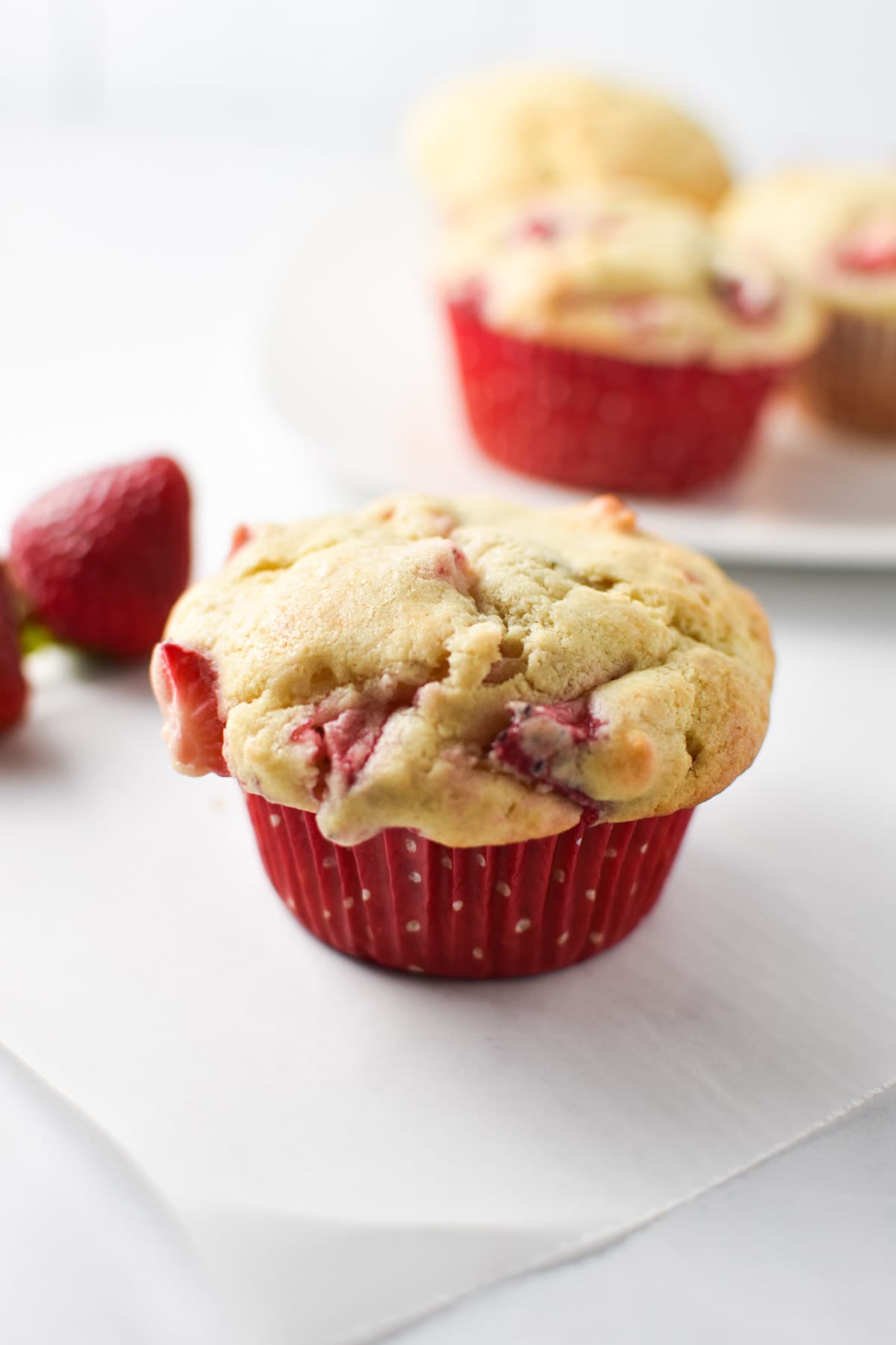 A muffin with strawberries on a white parchment paper surrounded by fresh strawberries.