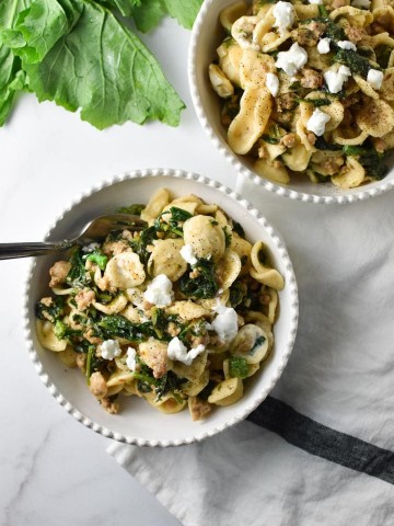 Two bowls of pasta with greens on the table next to a striped napkin with a fork