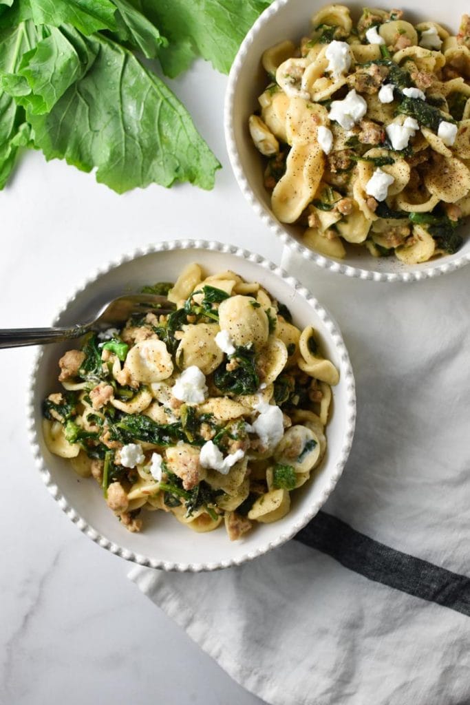 Two bowls of pasta with greens on the table next to a striped napkin with a fork