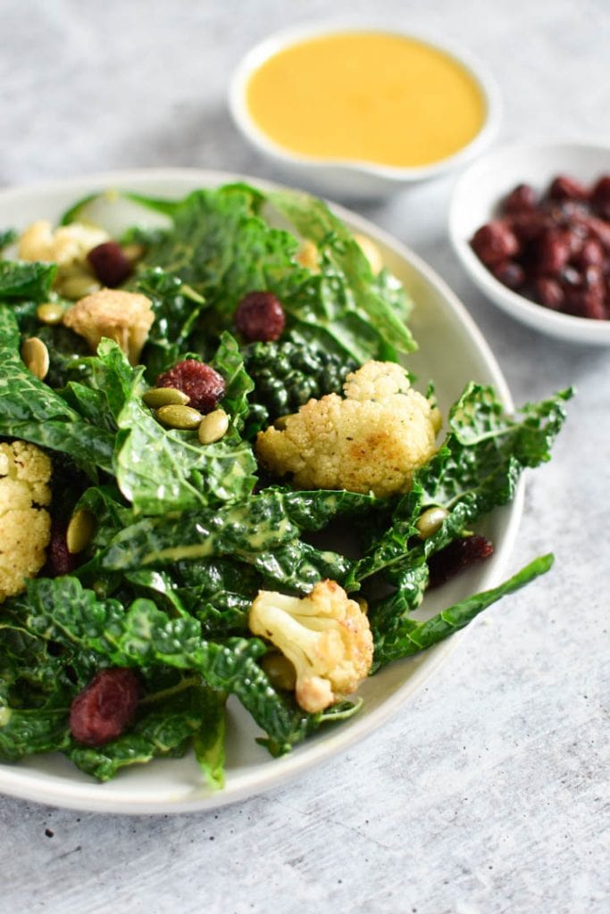 A kale salad topped with cauliflower next to dressing and cranberries