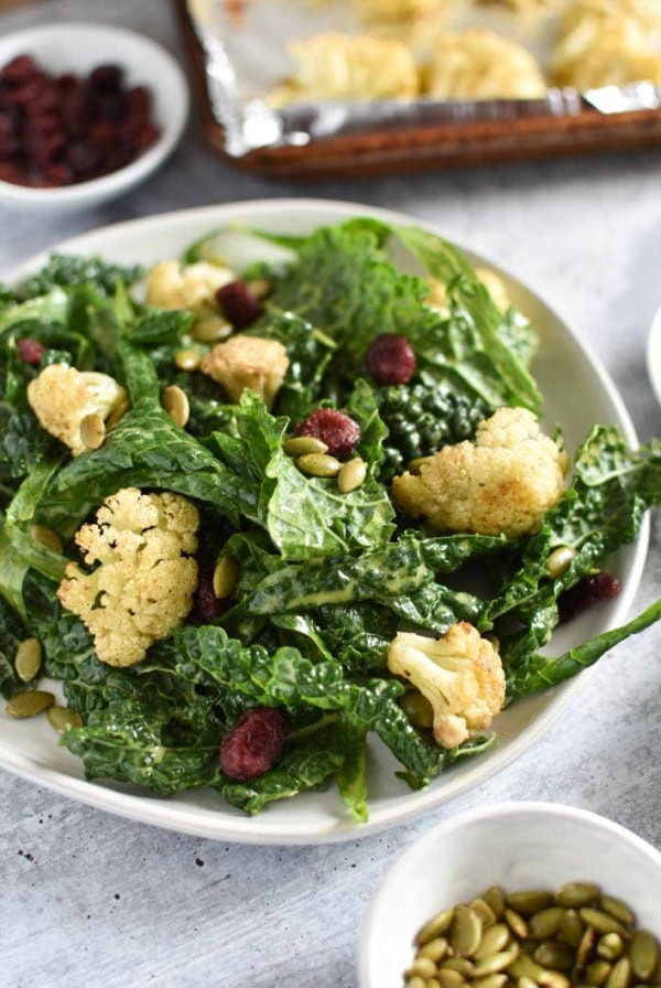 A bowl of kale cauliflower salad next to seeds and a baking sheet