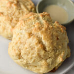 Golden brown drop biscuits piled on a gray plate next to a bowl of maple glaze