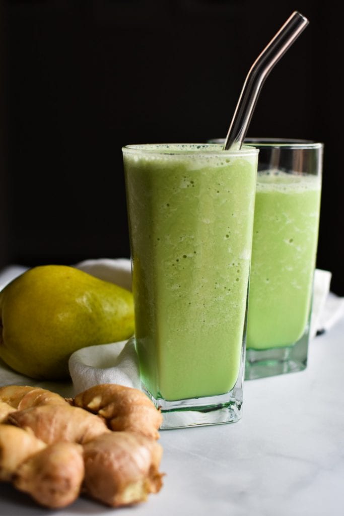 Two green smoothies with a glass straw next to a pear and ginger knob