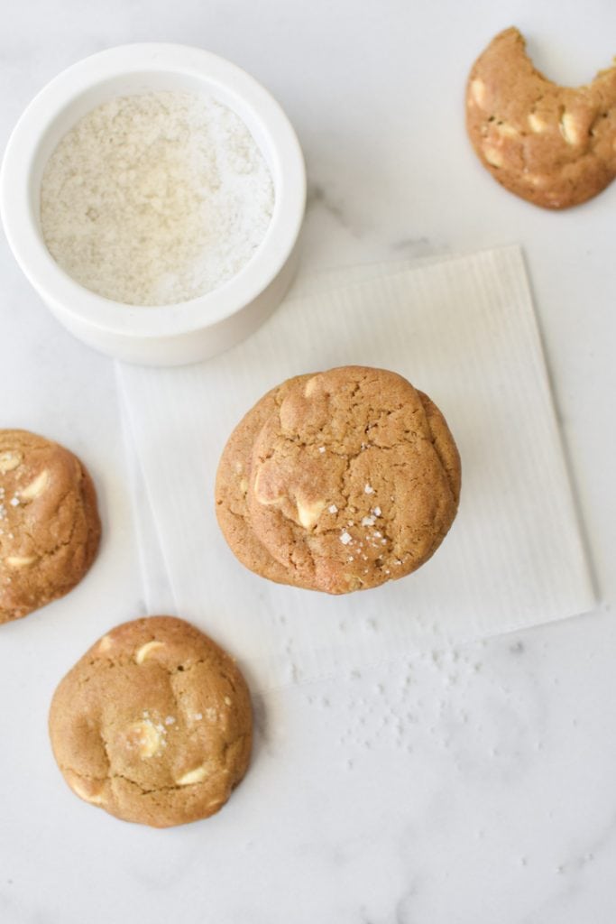 Stacked white chocolate chip cookies on a table, one with a bite out of it, next to a bowl of sea salt