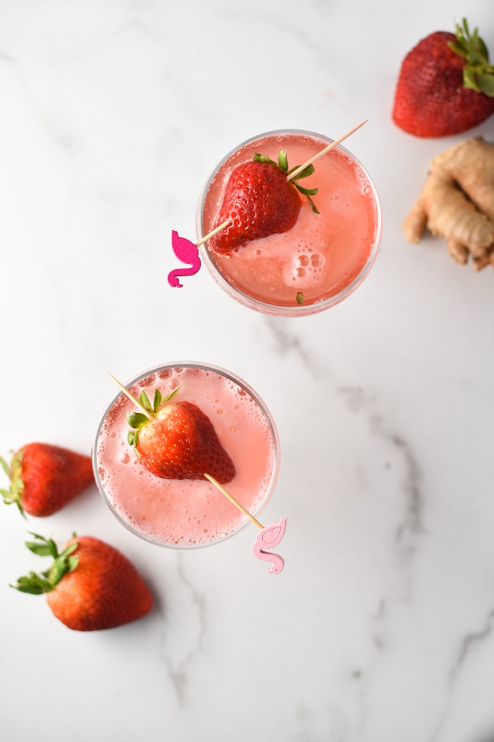 An overhead shot of two coupe glasses of strawberry simple syrup with strawberries and ginger surrounding it. 