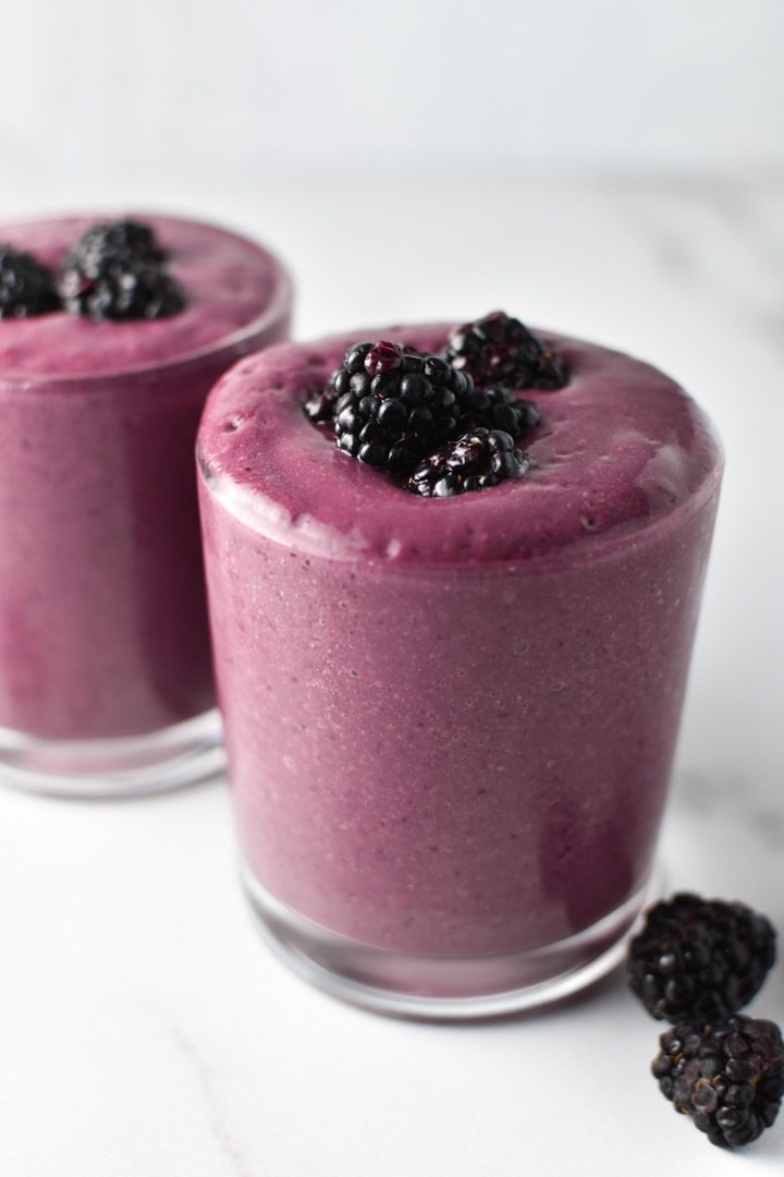 Two blackberry smoothies next to each other with fresh blackberries scattered next to the glasses