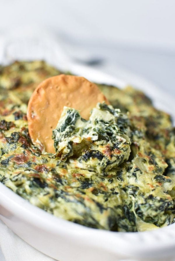 A round pita chip being dipped into a creamy spinach artichoke dip