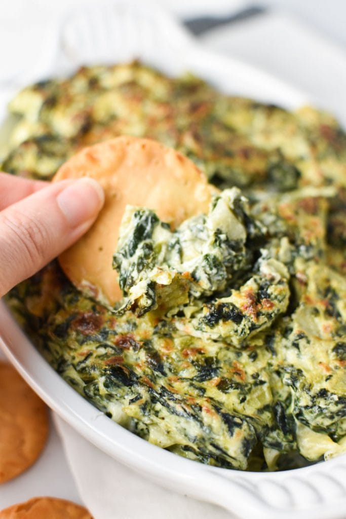 A hand dipping a pita chip into creamy spinach artichoke dip