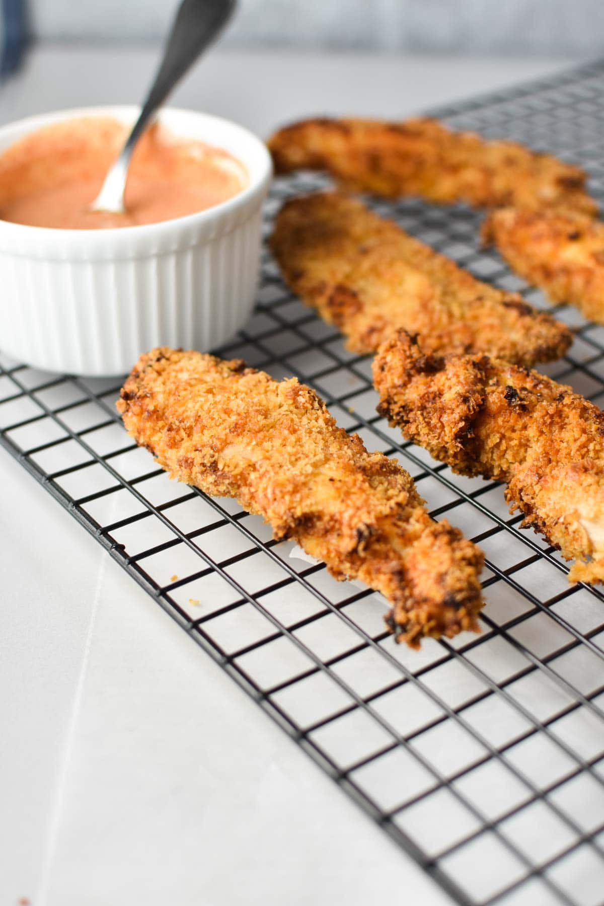 Air fried chicken tenders on a cooling rack next to a bowl of dipping sauce with a spoon. 