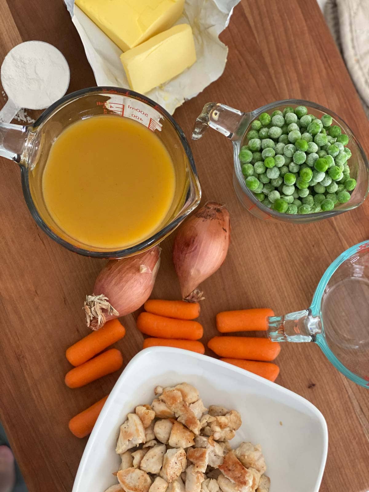 Broth, peas, carrots, shallots, chicken, butter, and milk on a wood board.