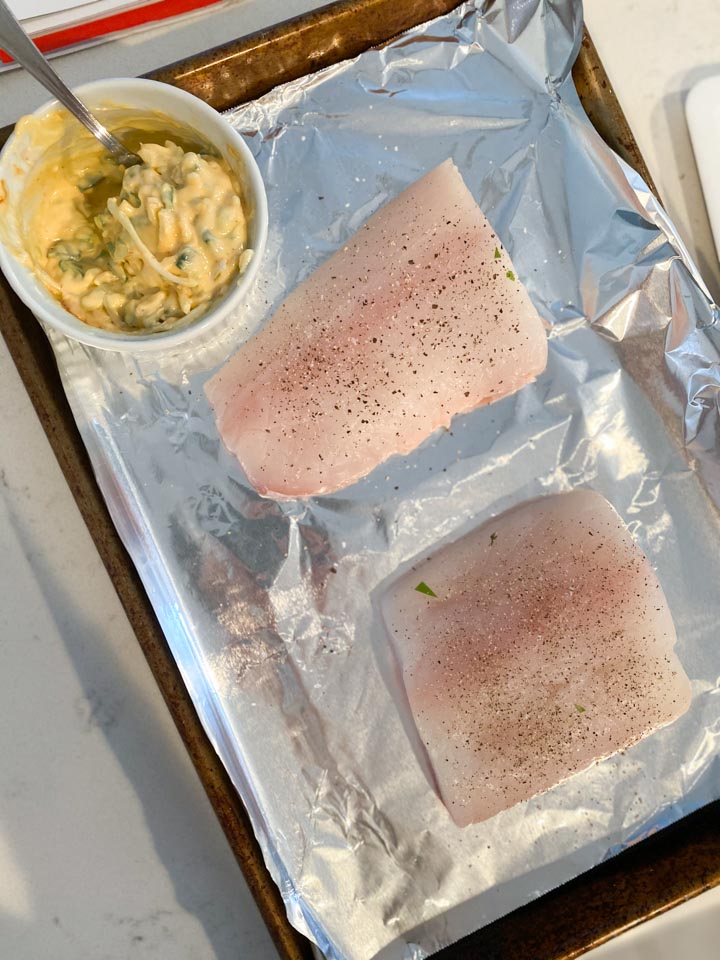 Two fresh halibut filets on a sheet pan next to a creamy sauce
