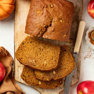 Slices of apple pumpkin bread on a serving platter next to a large loaf of the bread in the background.