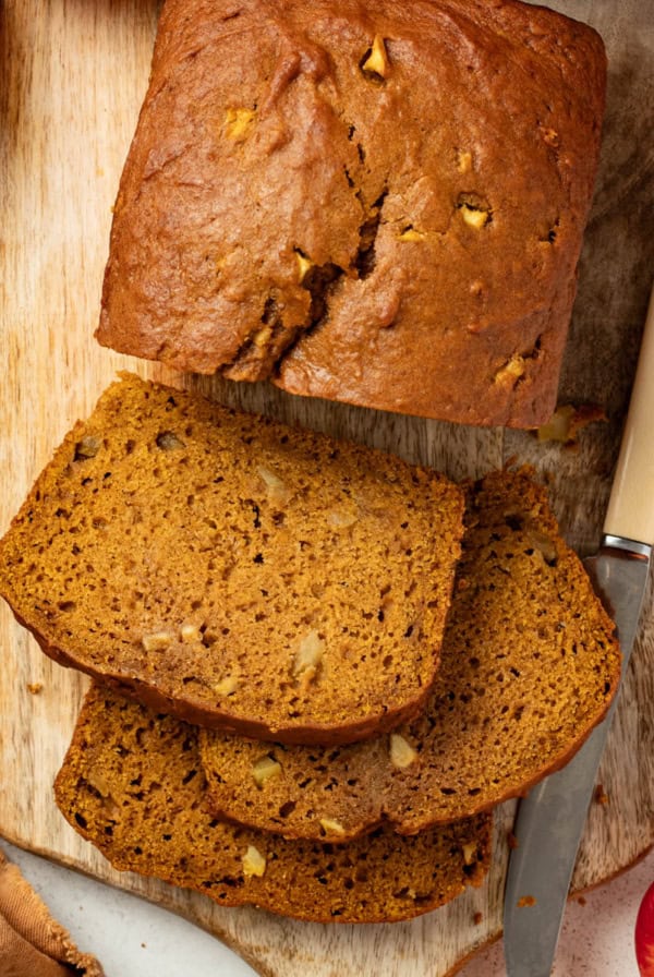 Slices of apple pumpkin bread on a serving platter next to a large loaf of the bread in the background.