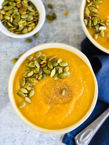 Two bowls of autumn squash soup next to a small bowl of pepitas