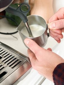 Hands frothing milk in a stainless steel steamer using a latte machine