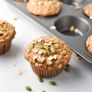 Cinnamon oatmeal muffins on a white table and in a baking pan, topped with oats and pumpkin seeds