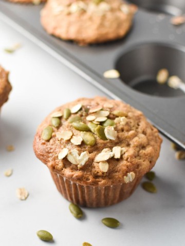 Cinnamon oatmeal muffins on a white table and in a baking pan, topped with oats and pumpkin seeds