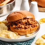 Sloppy joe sandwiches on a bun next to potato chips on a white plate.