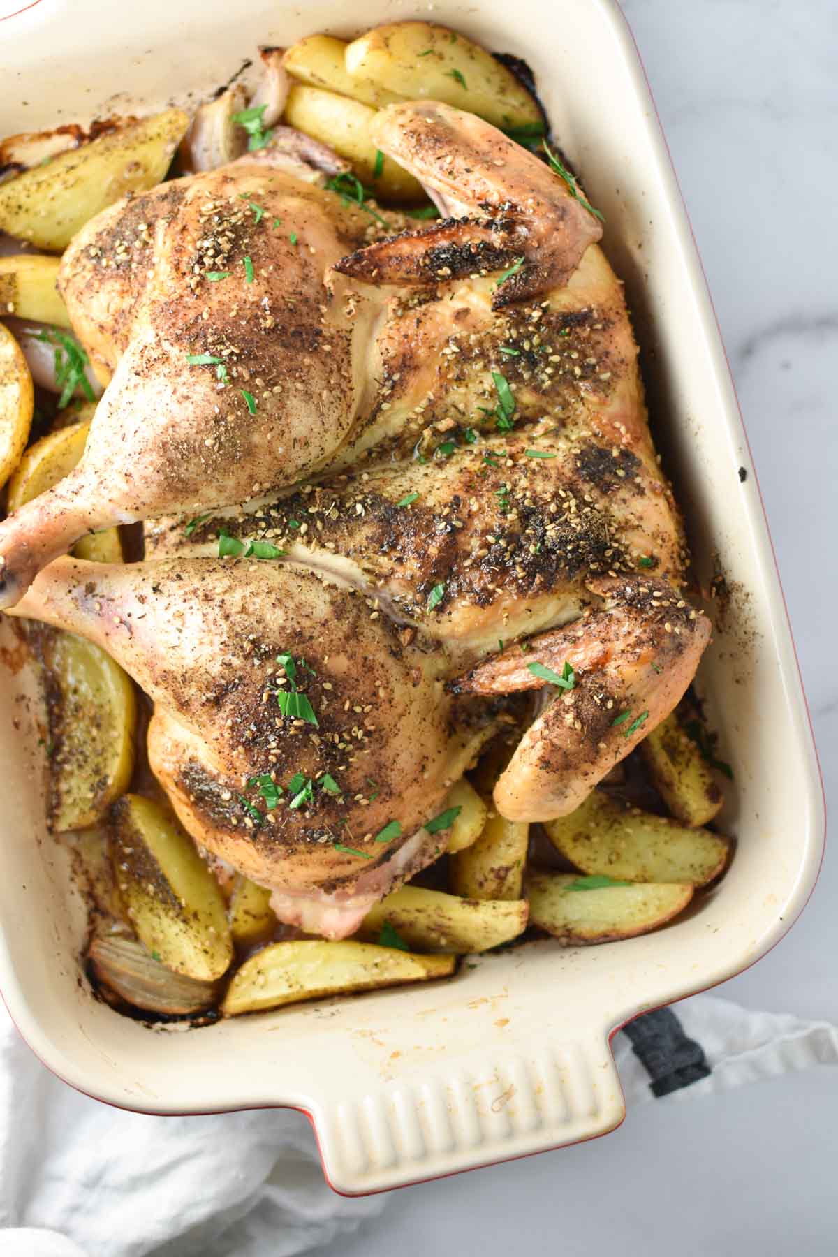 A roast chicken covered in za'atar and parsley in a baking dish with potatoes