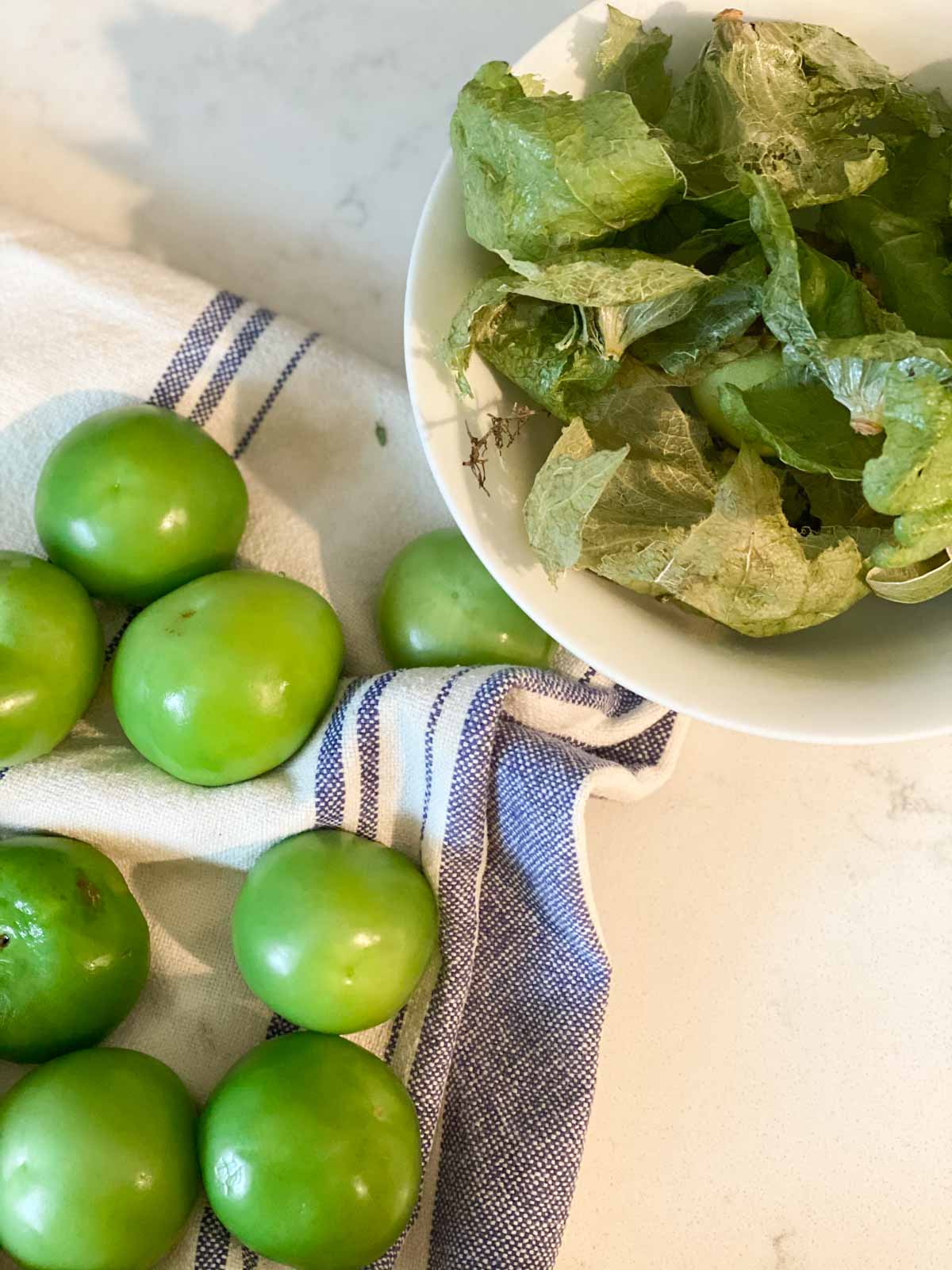 Peeled tomatillos on a towel with a bowl of their paper skins set aside