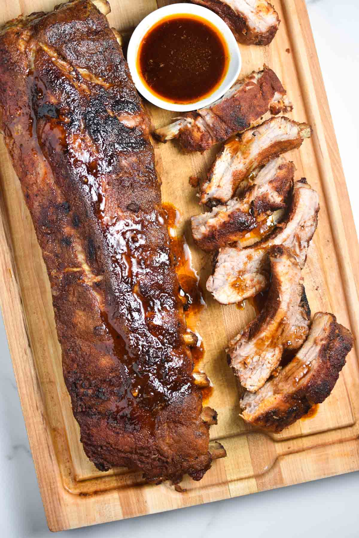 A whole rack of dry rub oven ribs next to sliced ribs and bbq sauce on a wooden board