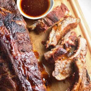 A whole rack of dry rubbed oven ribs next to sliced ribs and bbq sauce on a wooden board