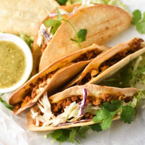 Baked chicken tacos on a white plate with slaw and a bowl of salsa verde