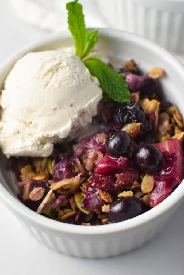 A strawberry blueberry crisp topped with oats and vanilla ice cream with a sprig of mint in a white bowl