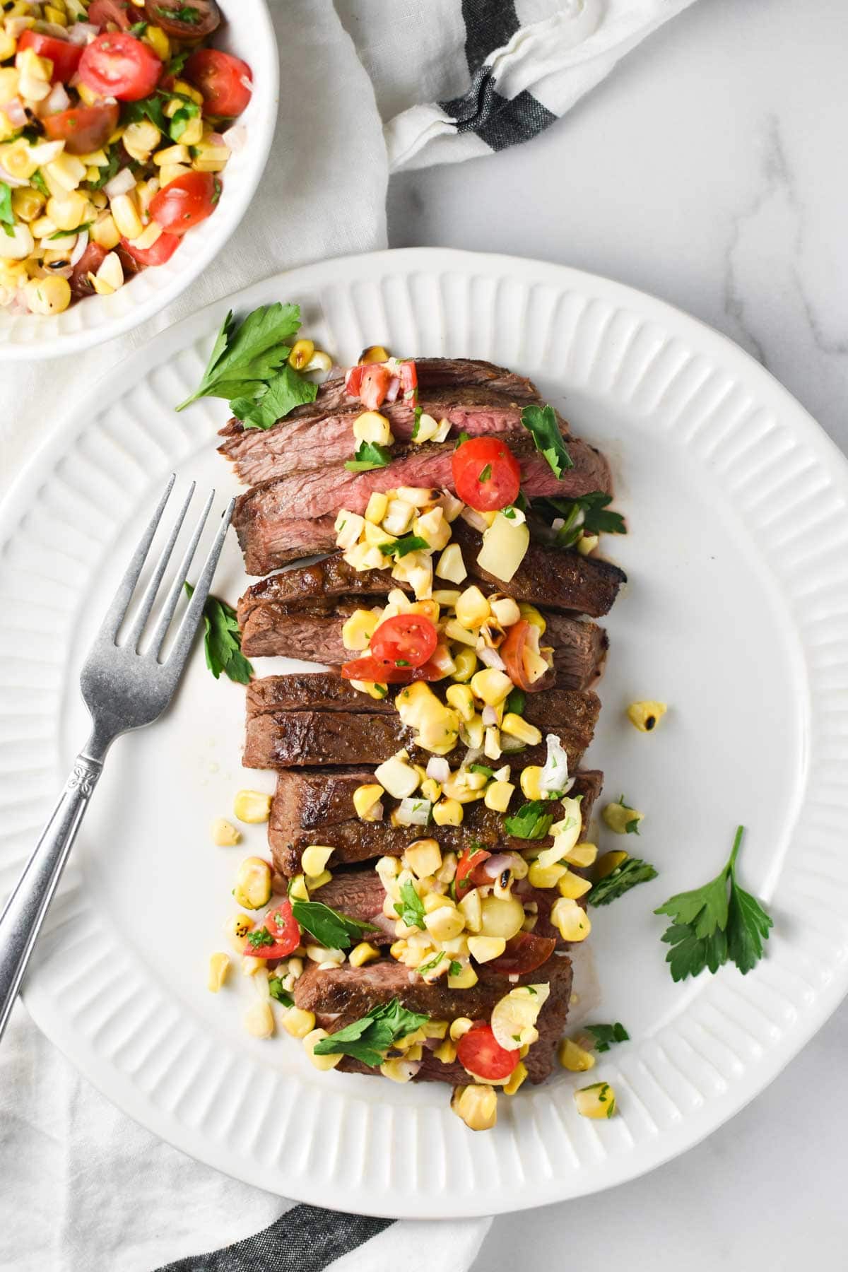 Sliced flank steak on a white plate topped with corn salsa and a fork next to a bowl of corn salsa