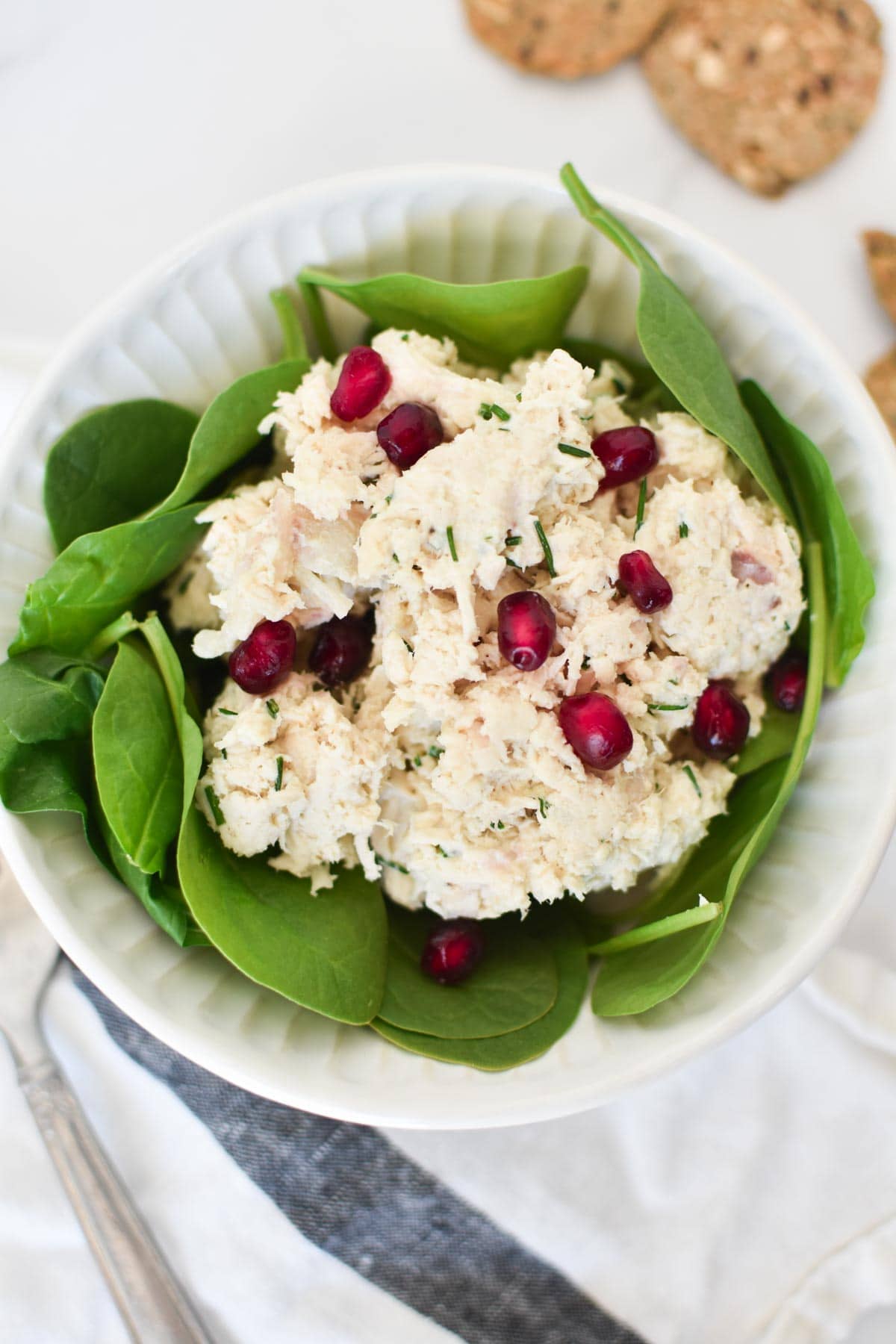 A pomegranate chicken salad in a white bowl next to a fork and crackers