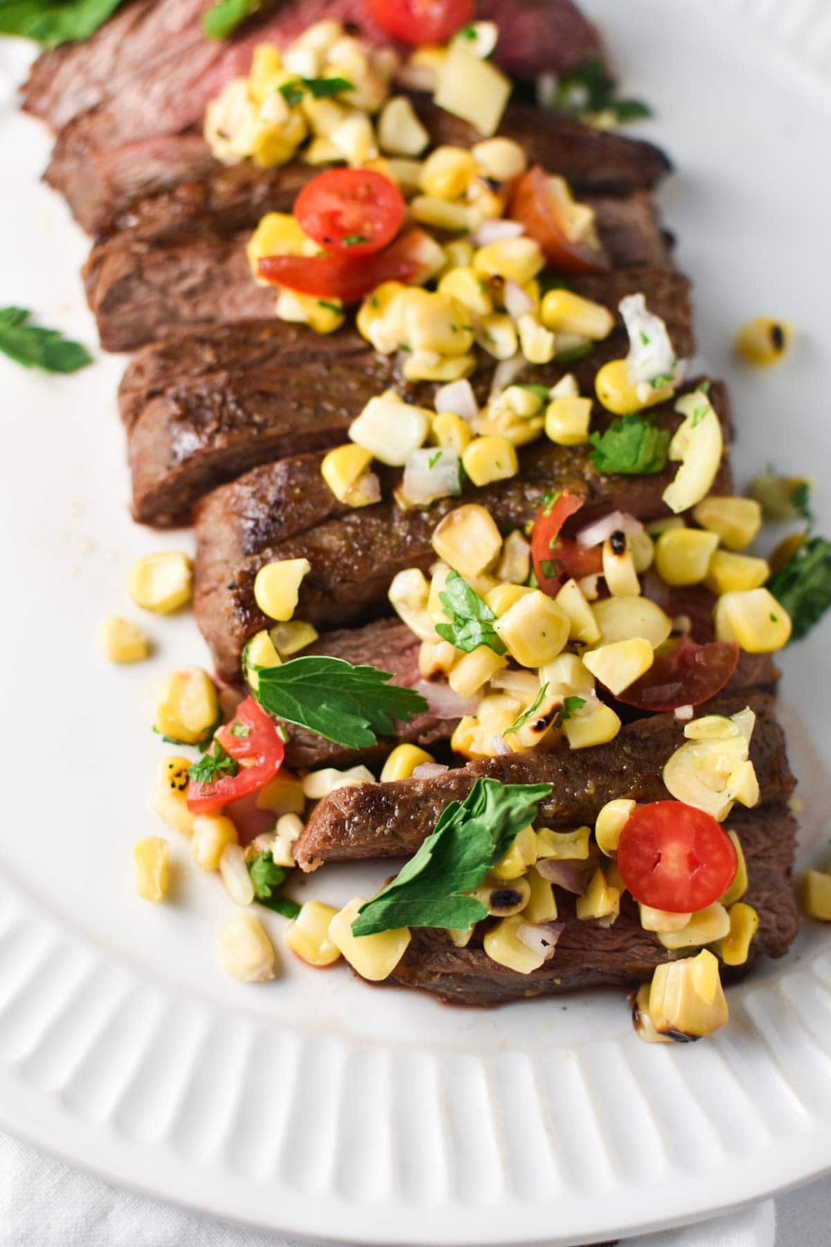 Sliced steak on a white plate topped with corn, tomatoes, and parsley