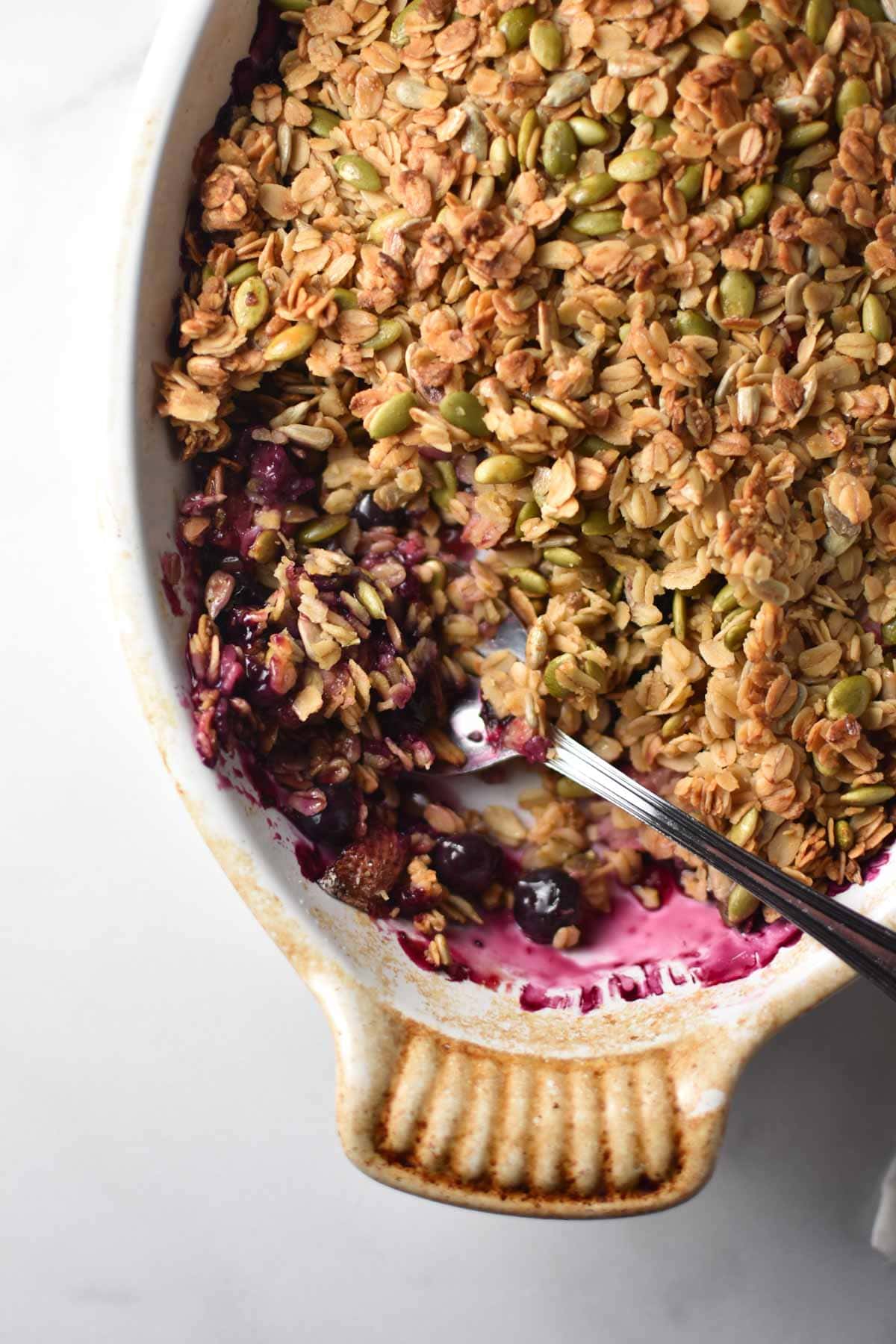 A spoon digging into a gratin dish with a berry crisp topped with seeds and oatmeal