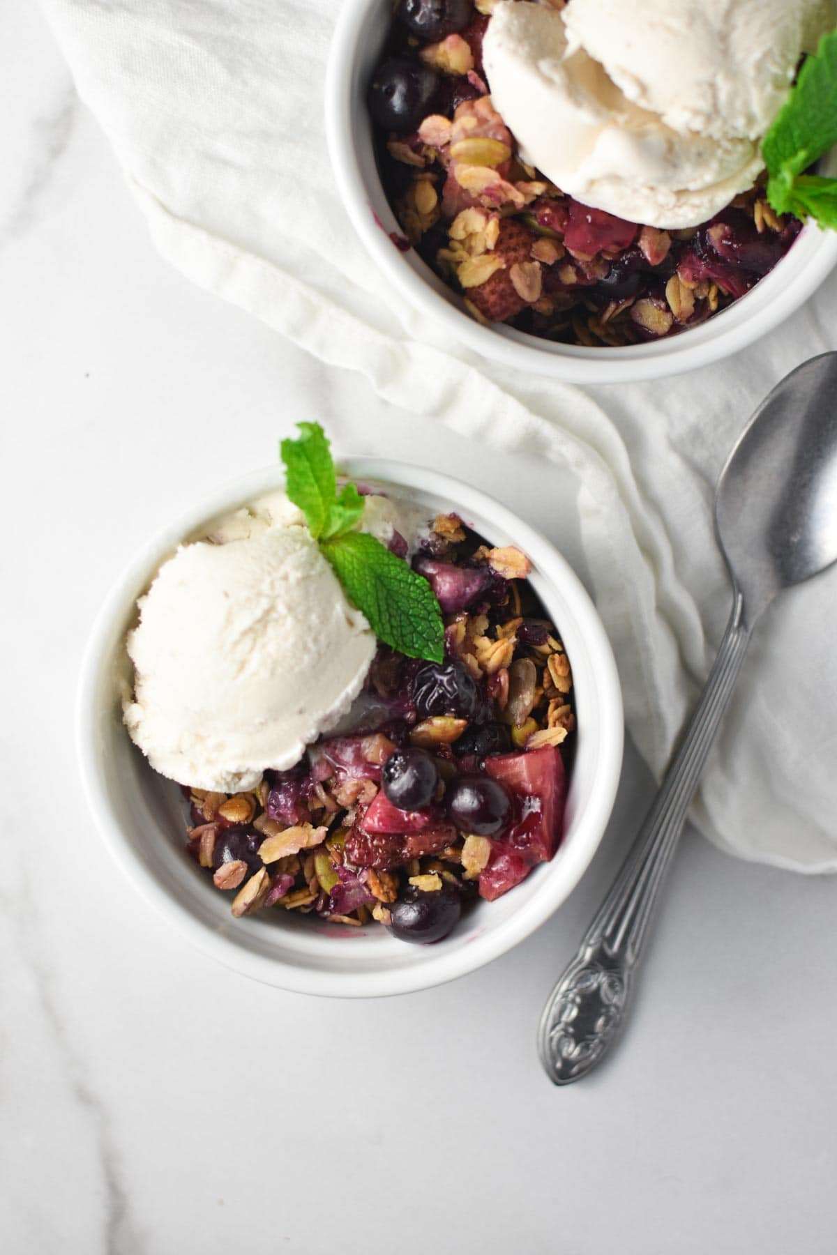 Two berry crisps with ice cream on top of a white marble table with a spoon