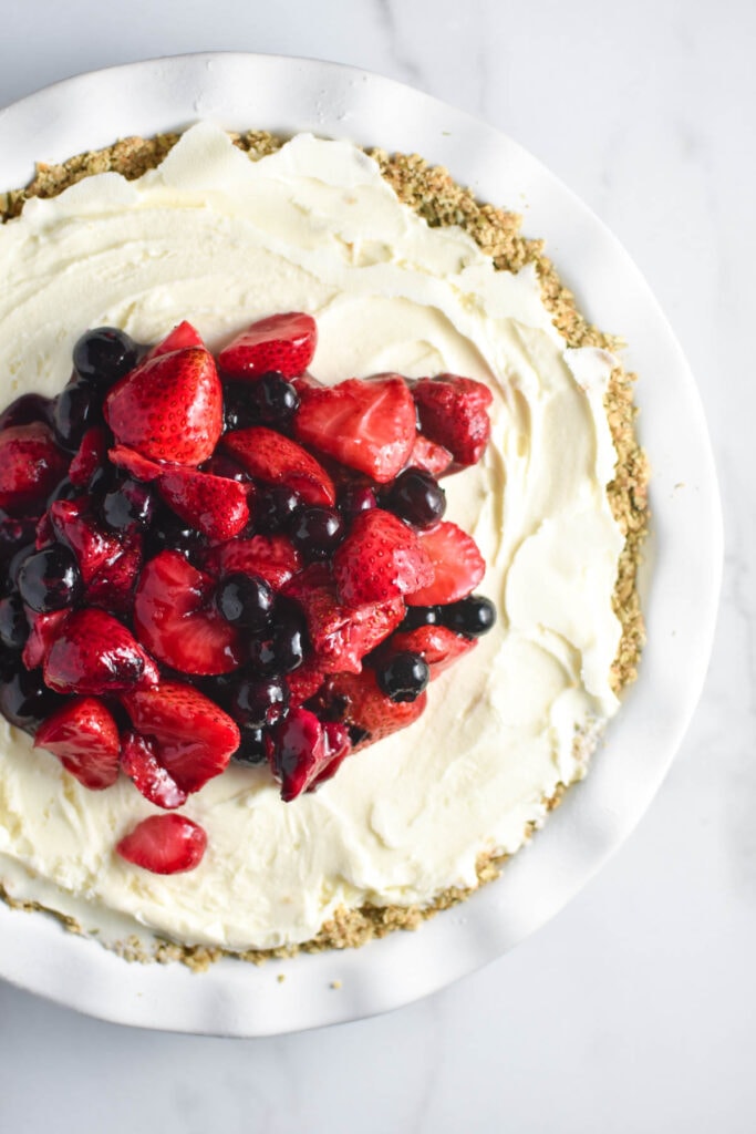 A vanilla ice cream pie on a white table topped with strawberries and blueberries