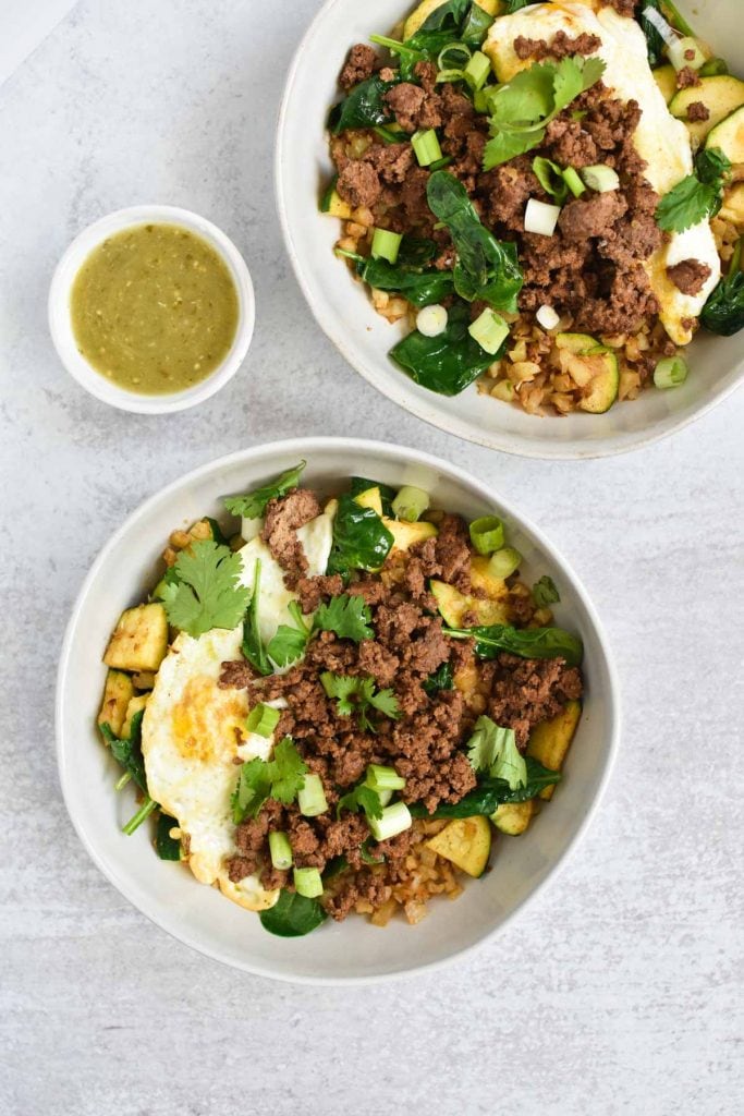 Two protein bowls filled with seasoned beef, eggs, and vegetables on a white table with salsa verde