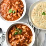 Two bowls of chicken ruby curry, one with cream on top, next to a plate of naan bread and a spoon and fork on a white table