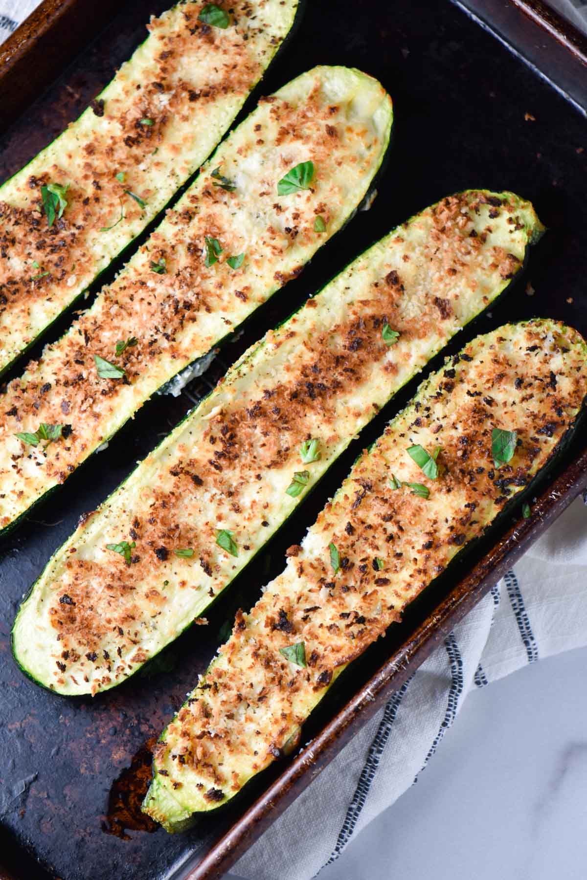 Four zucchini boats with a light brown panko crust on a metal sheet pan and towel